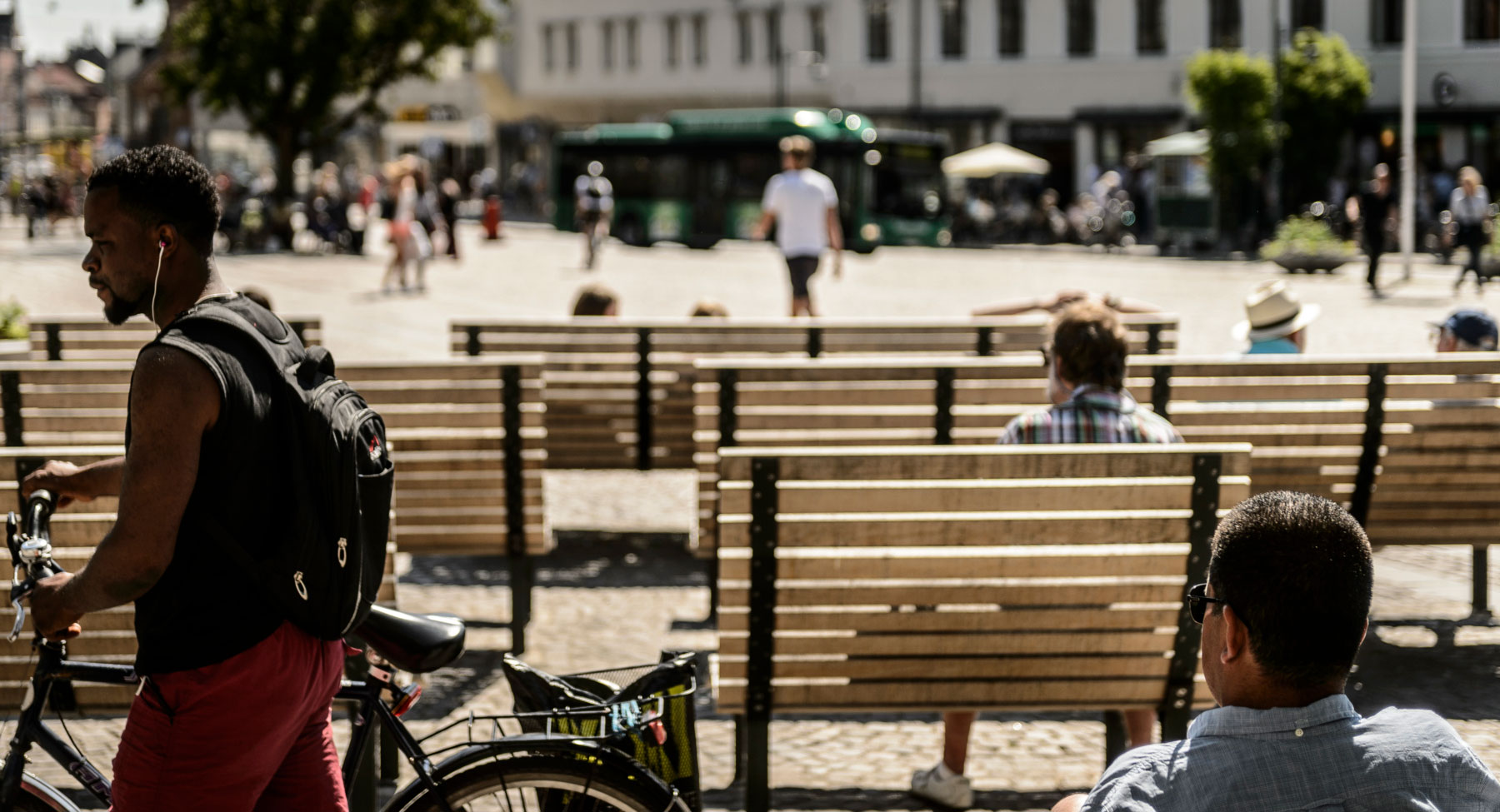 A man walking with his bike across Stortorget in Lund, where people are sitting at benches. Photo.